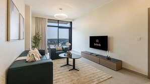 A modern living room with a sofa, a coffee table, a TV, and a large window offering a city view. - Brand-New 1BR in Neva Residence | Near Circle Mall (Dubai)