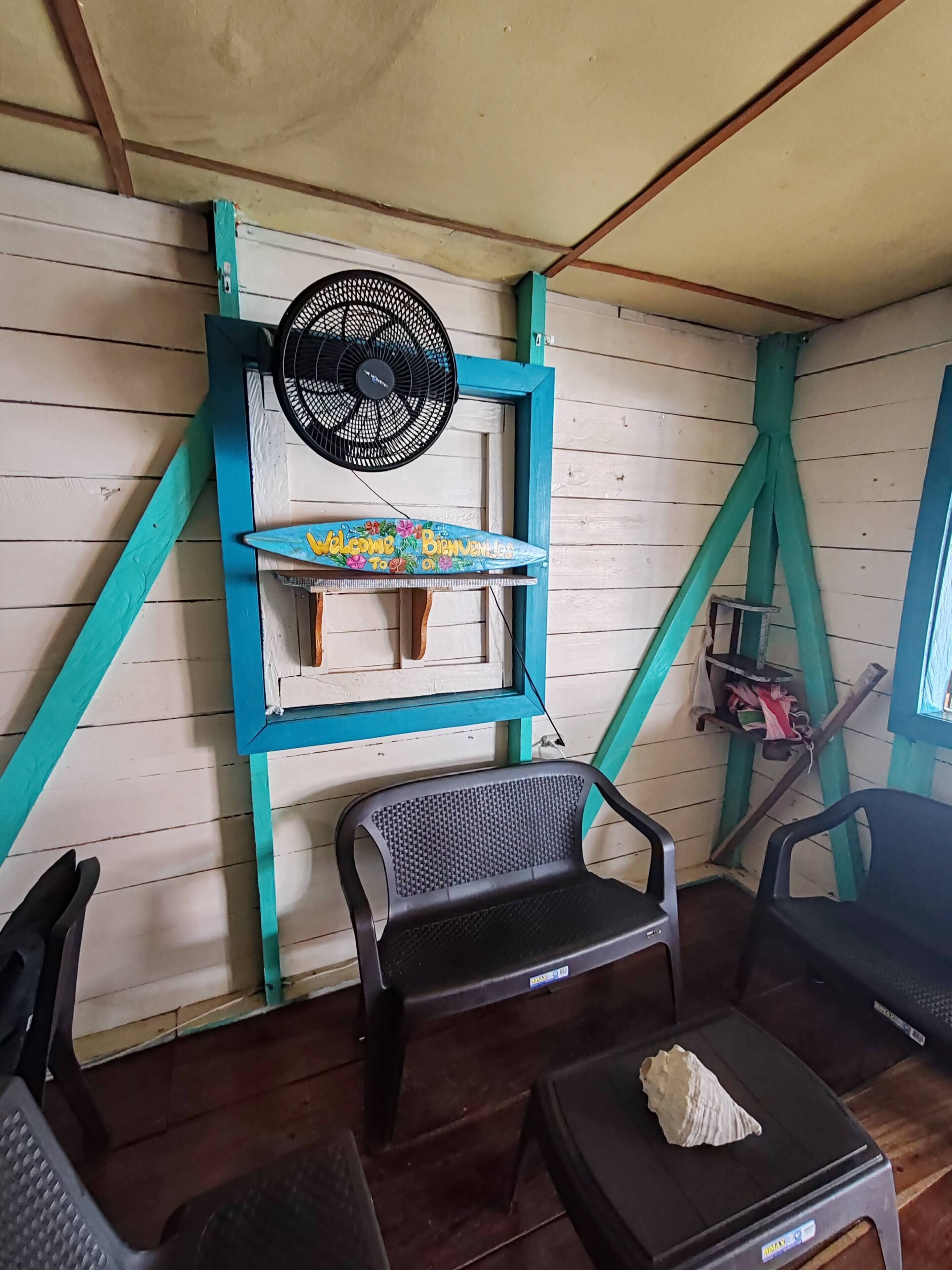 Una habitación con un ventilador en la pared, un estante con un letrero de bienvenida, dos sillas y una mesa pequeña con una concha encima.