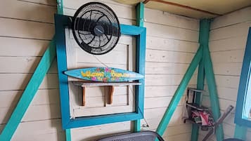 Una habitación con un ventilador en la pared, un estante con un letrero de bienvenida, dos sillas y una mesa pequeña con una concha encima.