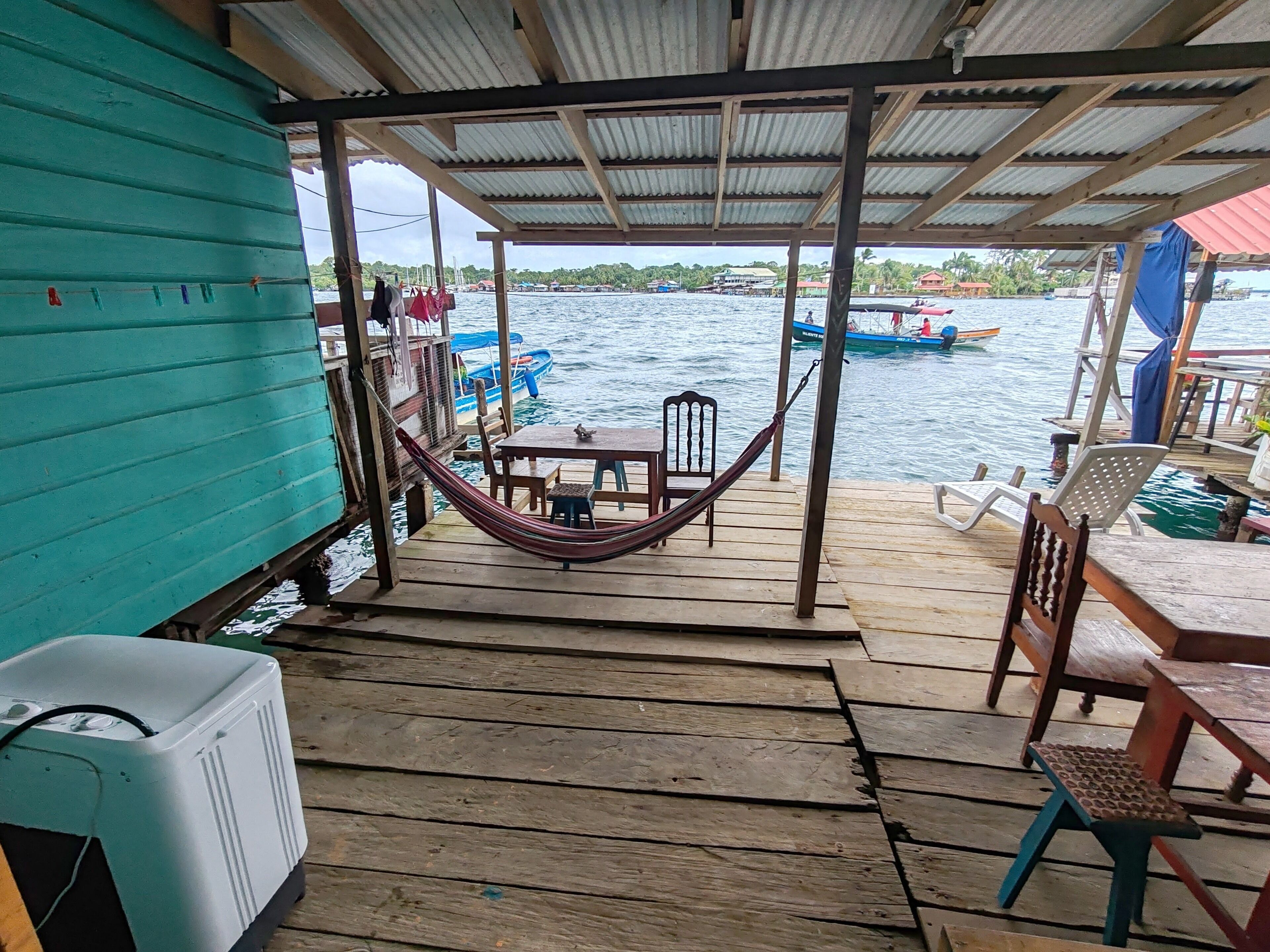Un muelle de madera con una hamaca, una mesa y sillas, con vista a un cuerpo de agua con barcos.