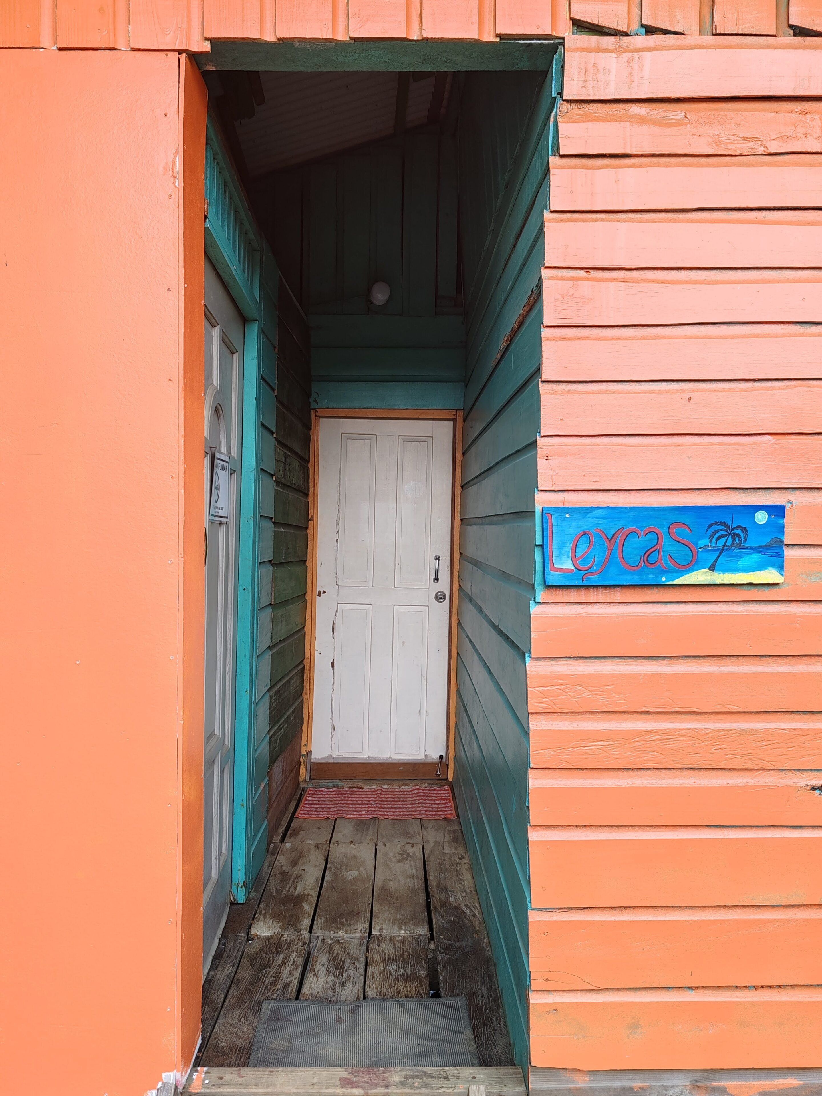 Une étroite entrée en bois avec une porte blanche, une enseigne bleue sur laquelle est écrit le mot « LOVAS », et un paillasson rouge au bas de l’entrée.