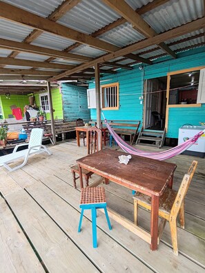 Una terraza de madera con una mesa, sillas, una hamaca y una pequeña mesa con mantel de encaje.