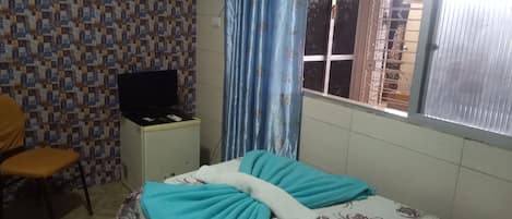 A bedroom with a bed, a television on a small cabinet, a chair, and a window with blinds.