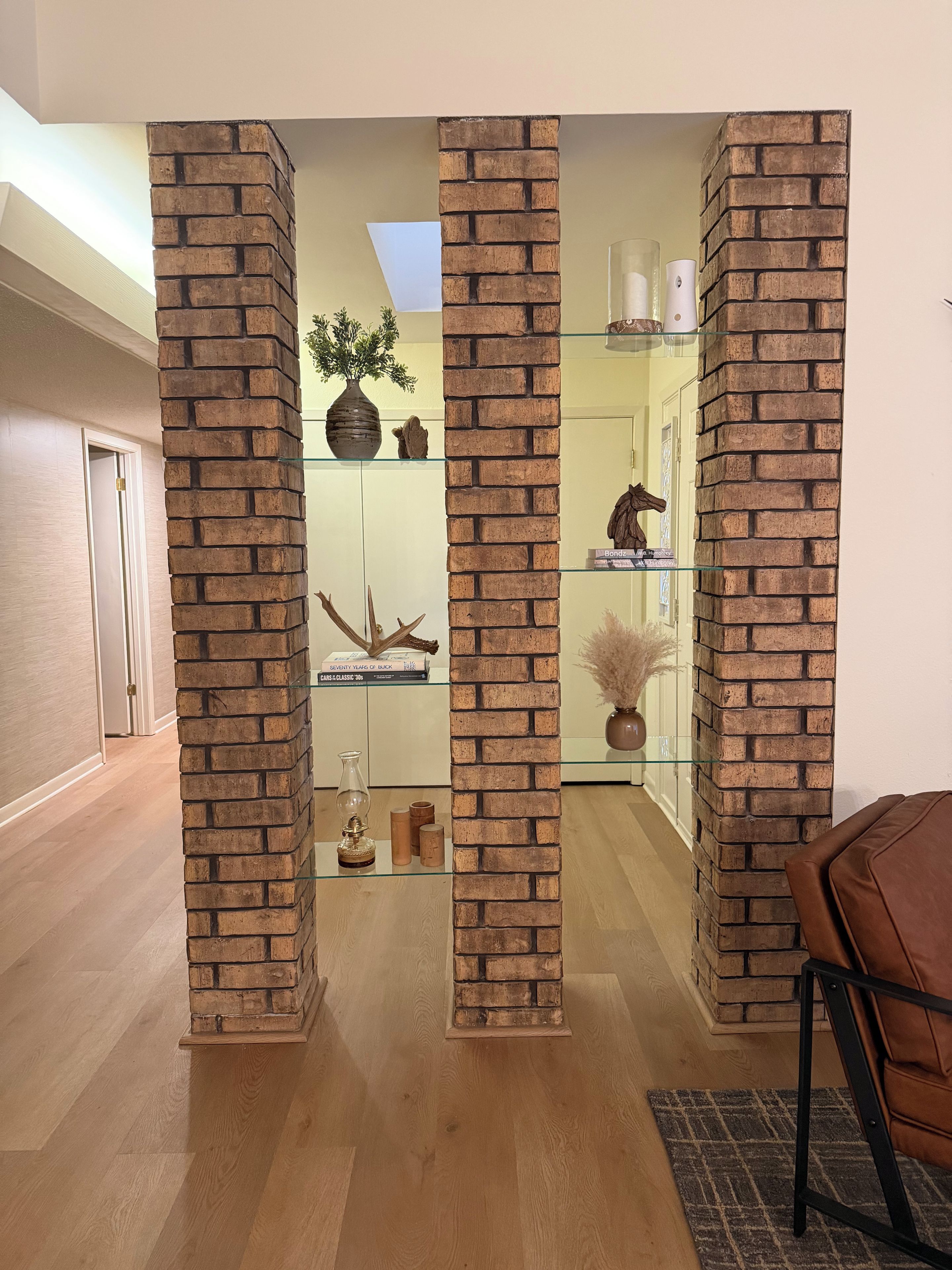 A room with a brick column structure, wooden flooring, a glass shelf with decorative items, and a brown leather chair.