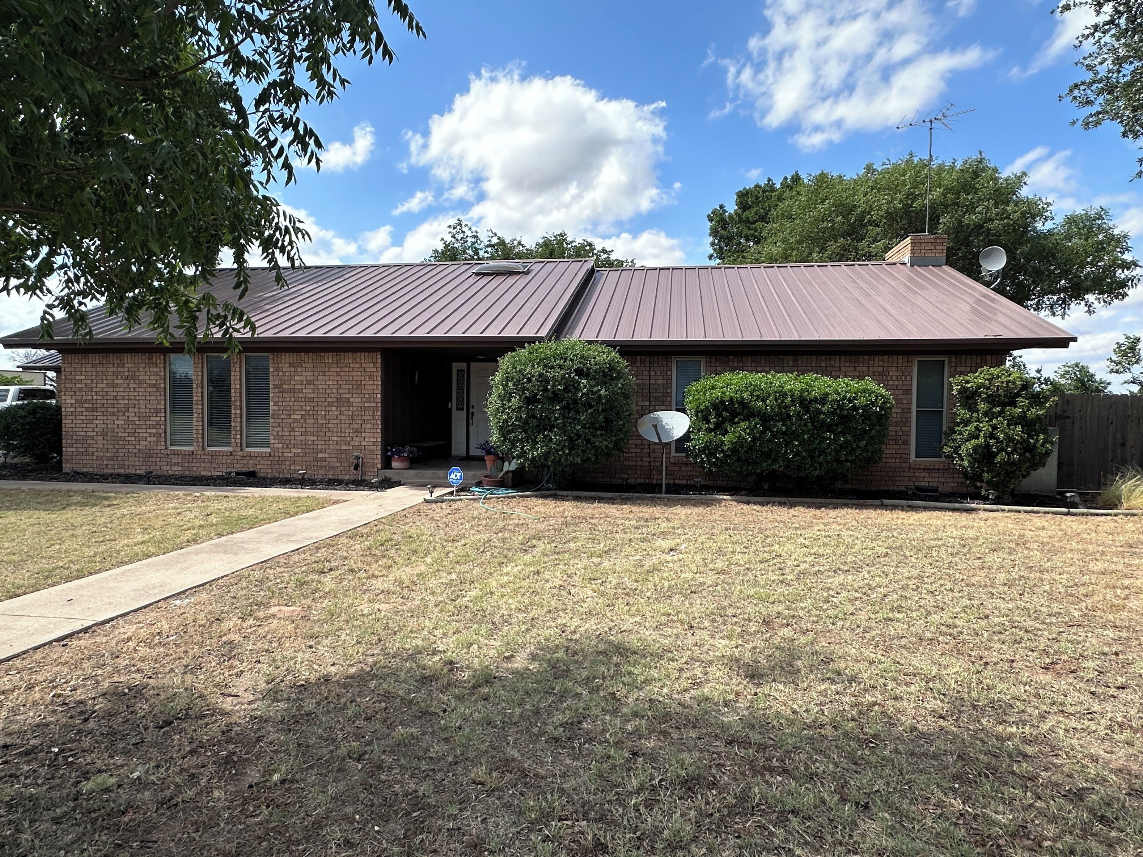 A single-story brick house with a metal roof, a front door, and a satellite dish.