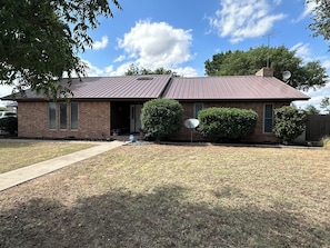 A single-story brick house with a metal roof, a front door, and a satellite dish.