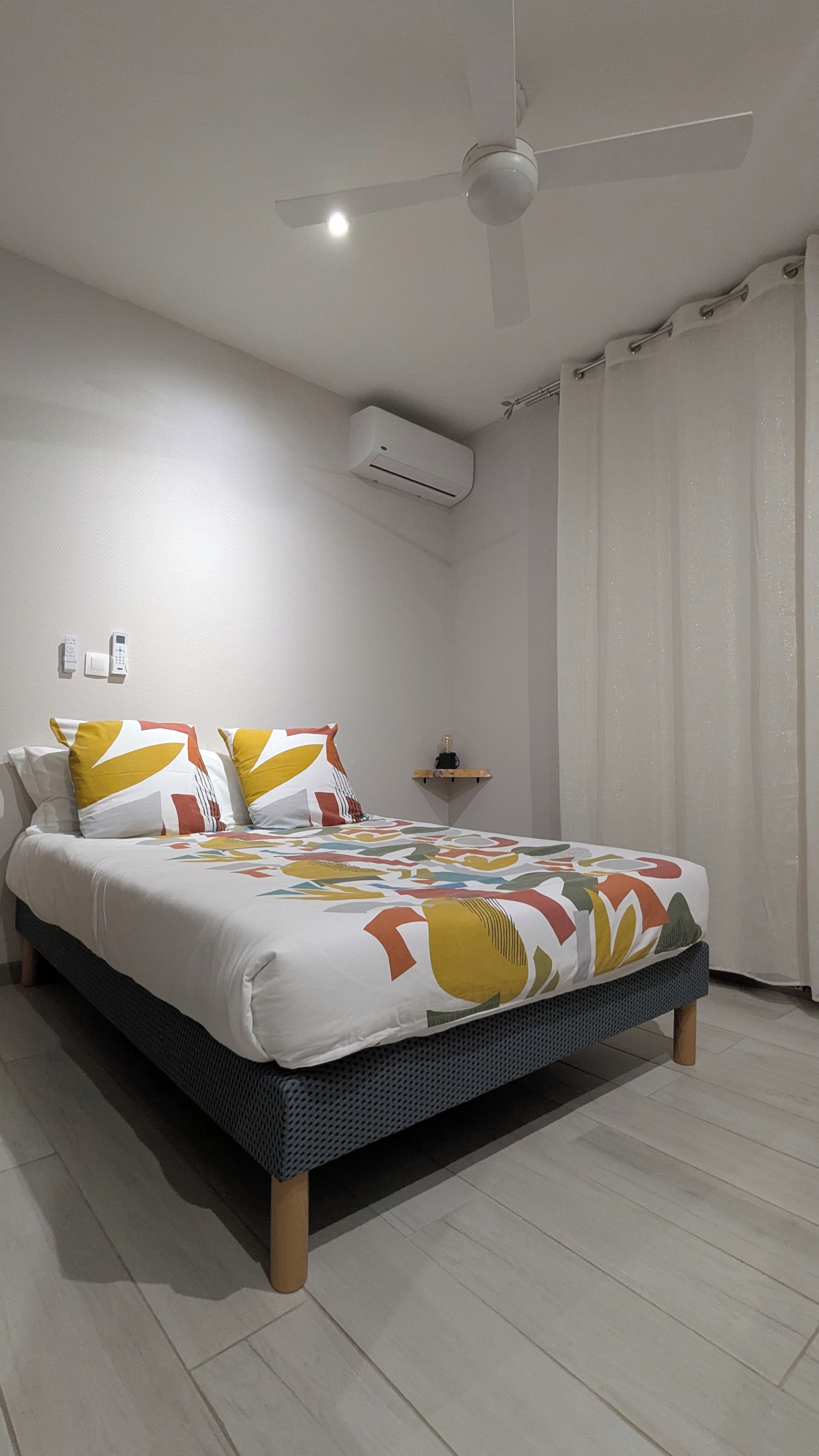 Une chambre à coucher comprenant un lit, un ventilateur de plafond, un appareil de climatisation, une table de chevet et une fenêtre avec des rideaux.