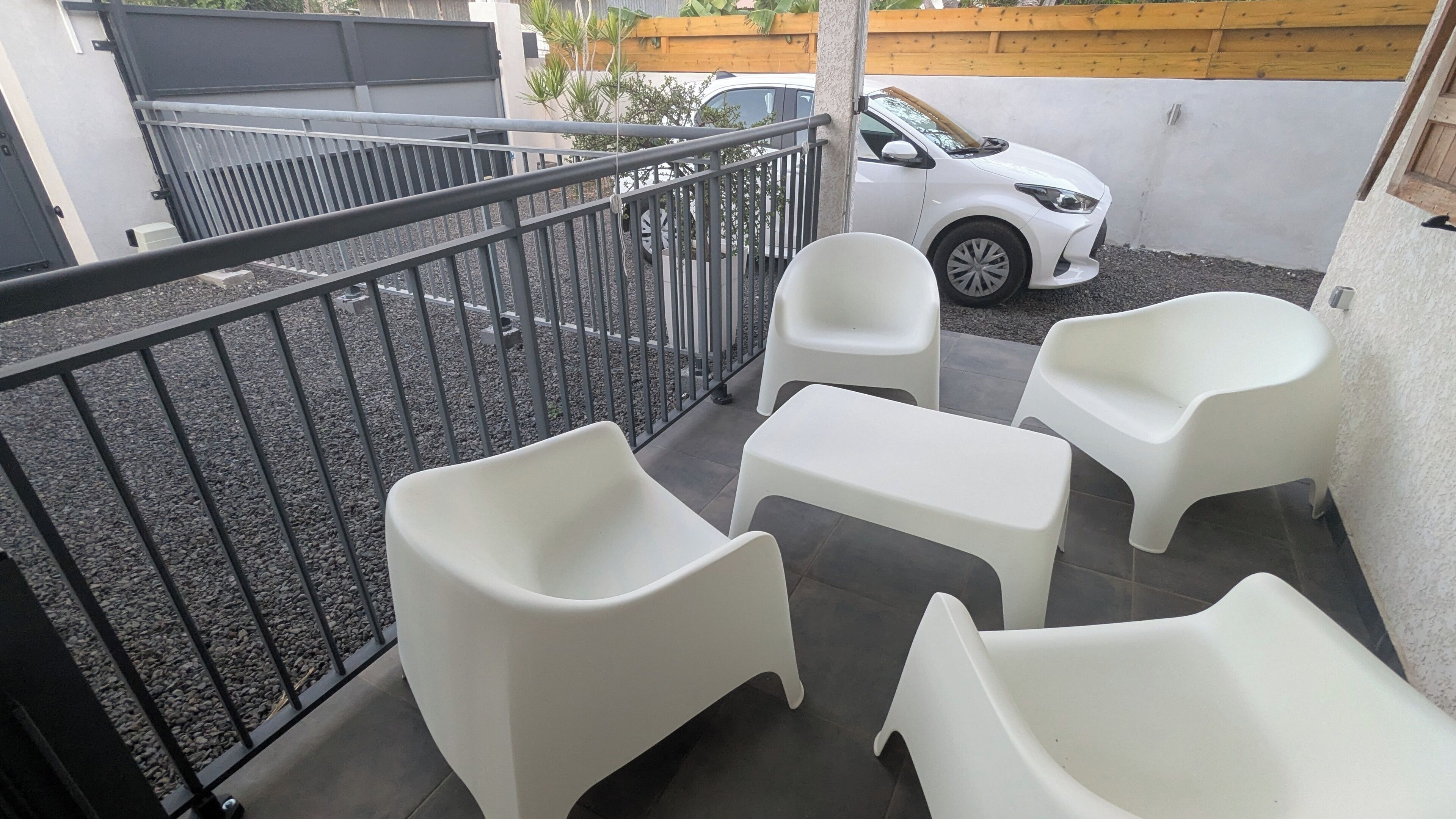 Un balcon avec une rambarde en métal, des chaises en plastique blanches et une voiture blanche garée à l’arrière-plan.