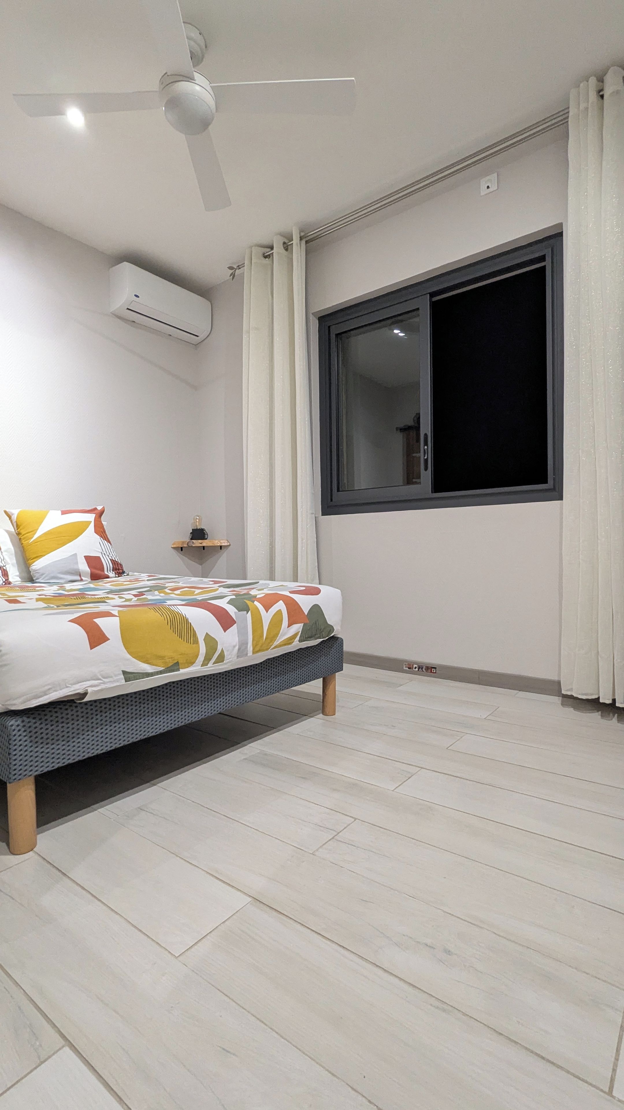 A bedroom with a bed, a ceiling fan, an air conditioning unit, a window with curtains, and a wooden floor.