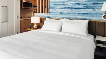 A hotel room with a large bed, a nightstand with a lamp, a wooden headboard, a painting on the wall, and a kitchenette with a microwave.