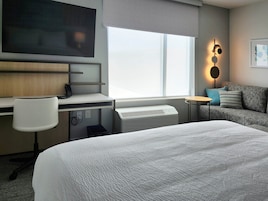 Habitación de hotel con cama, escritorio con silla, televisor, calentador, sofá con cojines y ventana con persianas.