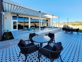 Un patio exterior con área de estar cubierta, piscina y un edificio con grandes ventanales.