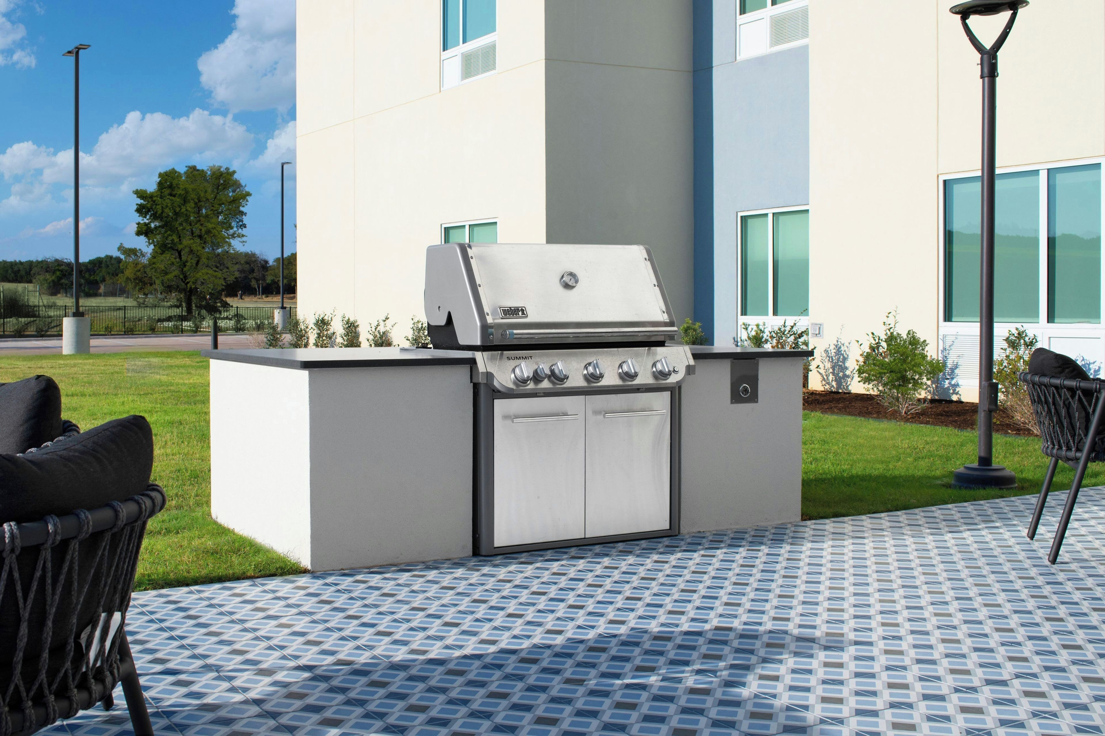 An outdoor grill station with a built-in oven, surrounded by a patterned patio floor, and adjacent to a modern building with large windows.
