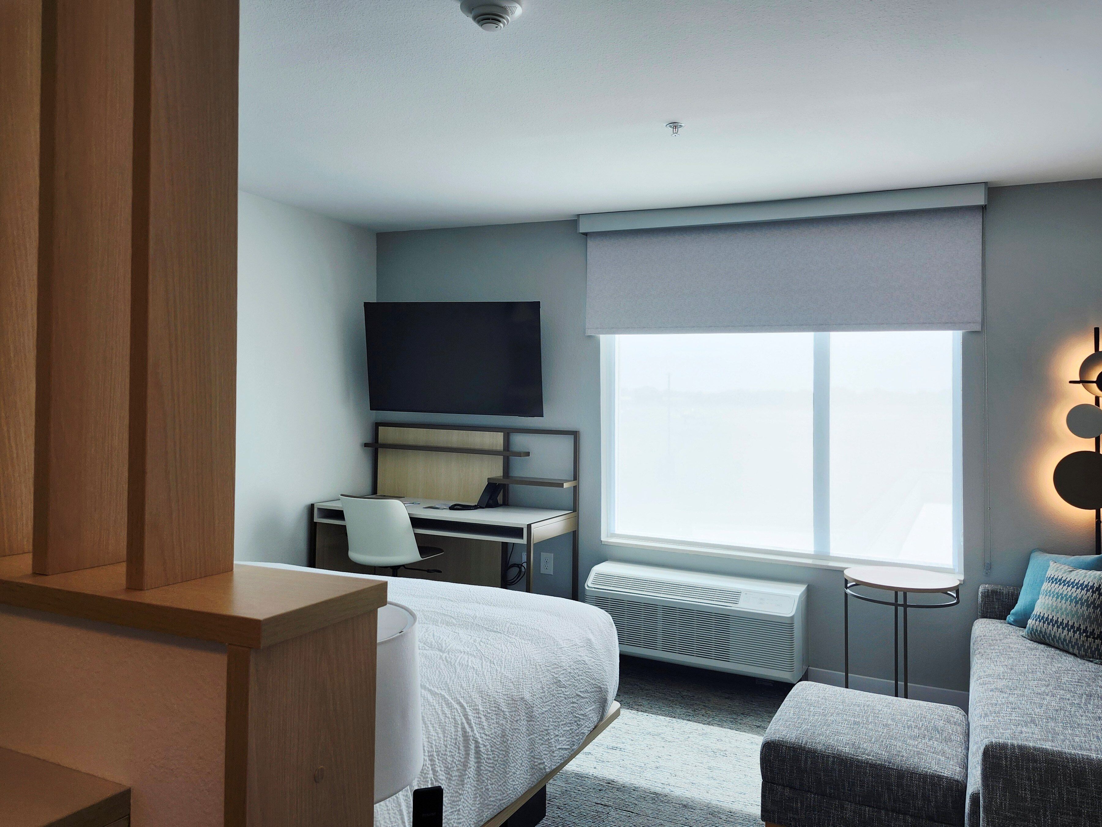 Habitación de hotel moderna con cama, escritorio con silla, televisor de pantalla plana, ventana con persianas, calefactor y una mesita.