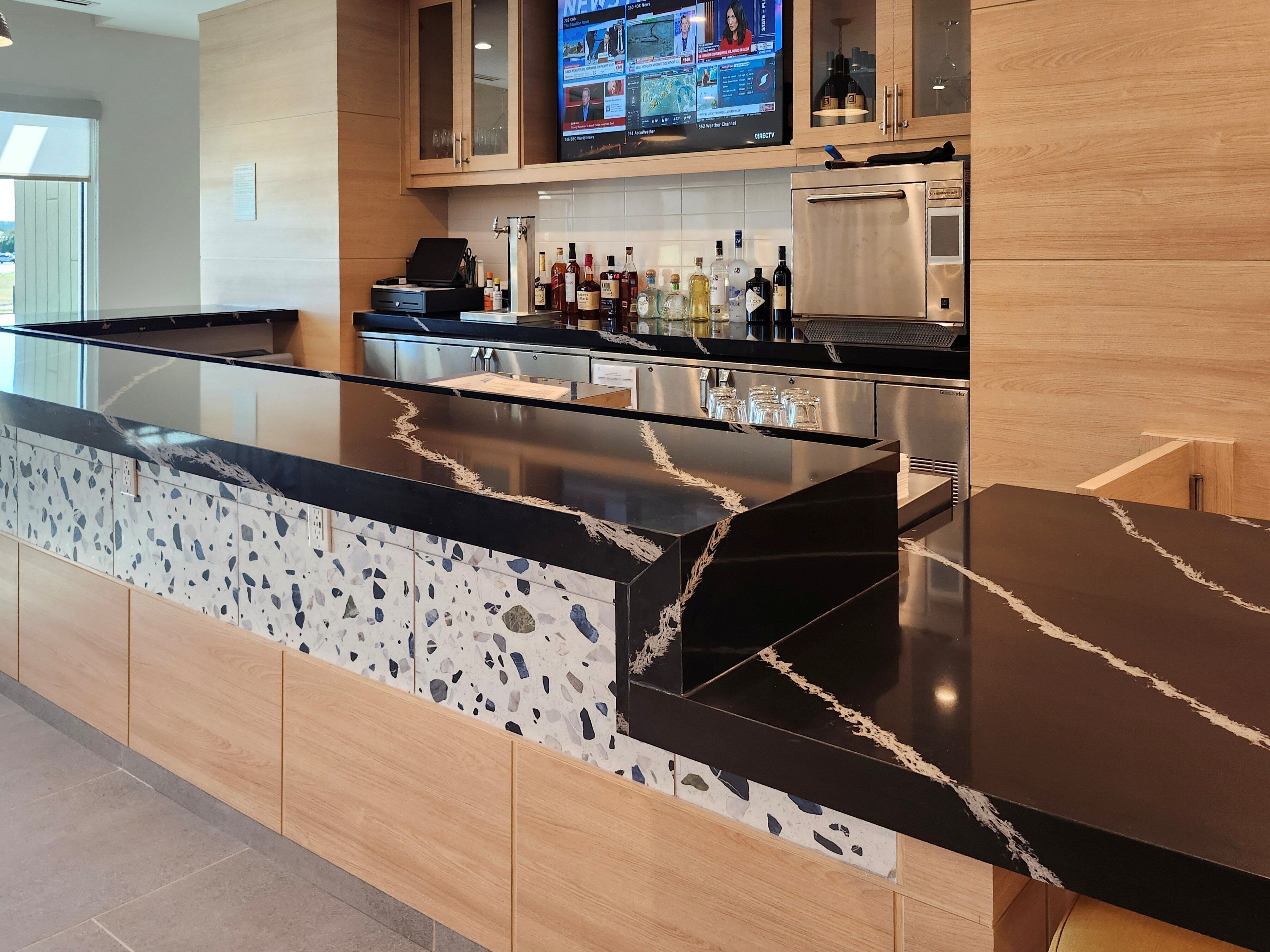 A modern bar area with a granite countertop, wooden cabinetry, and a flat-screen TV displaying news.