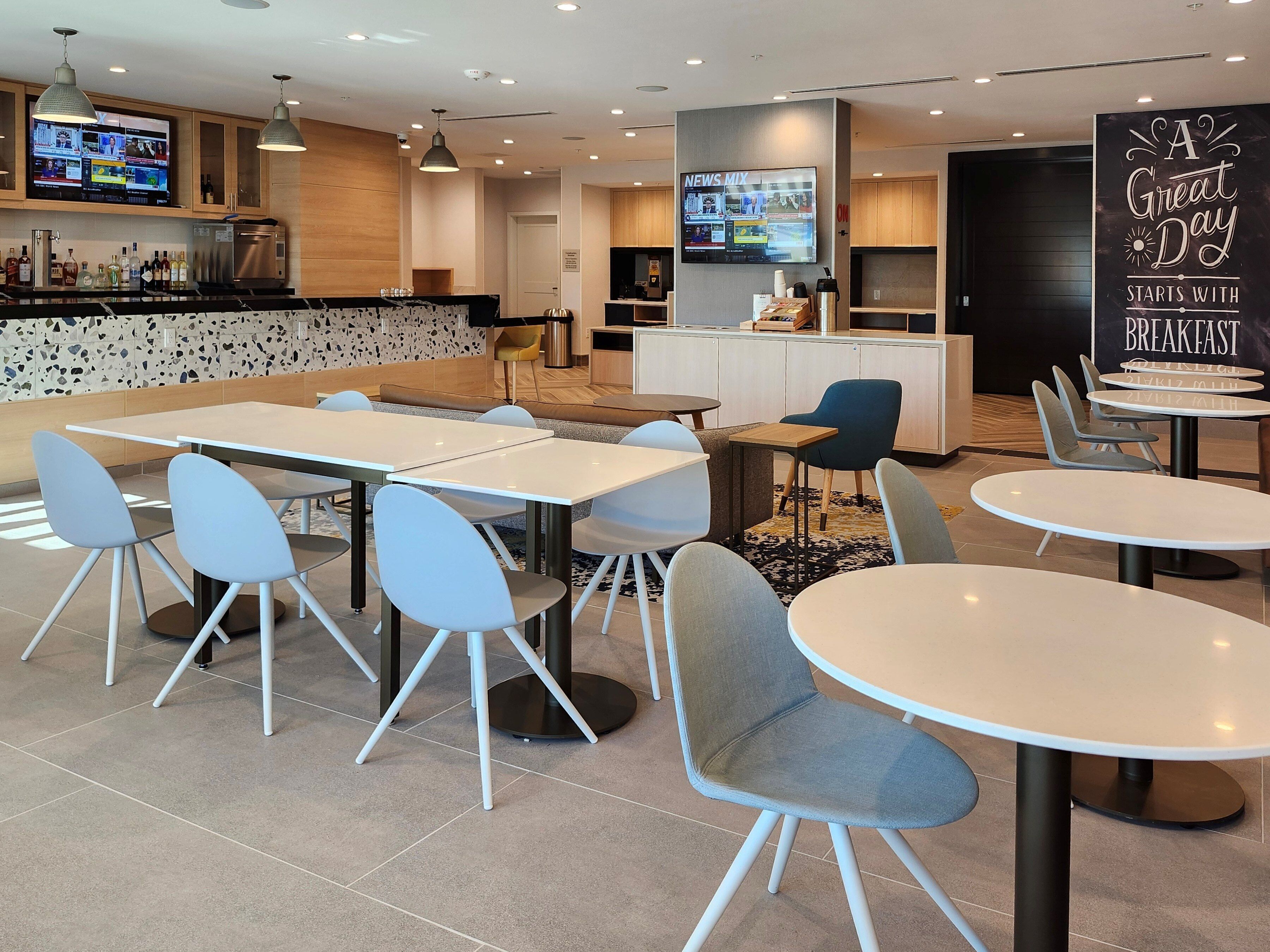 Un comedor moderno con varias mesas y sillas, una zona de cocina con un televisor, y una pared con una cita motivacional.