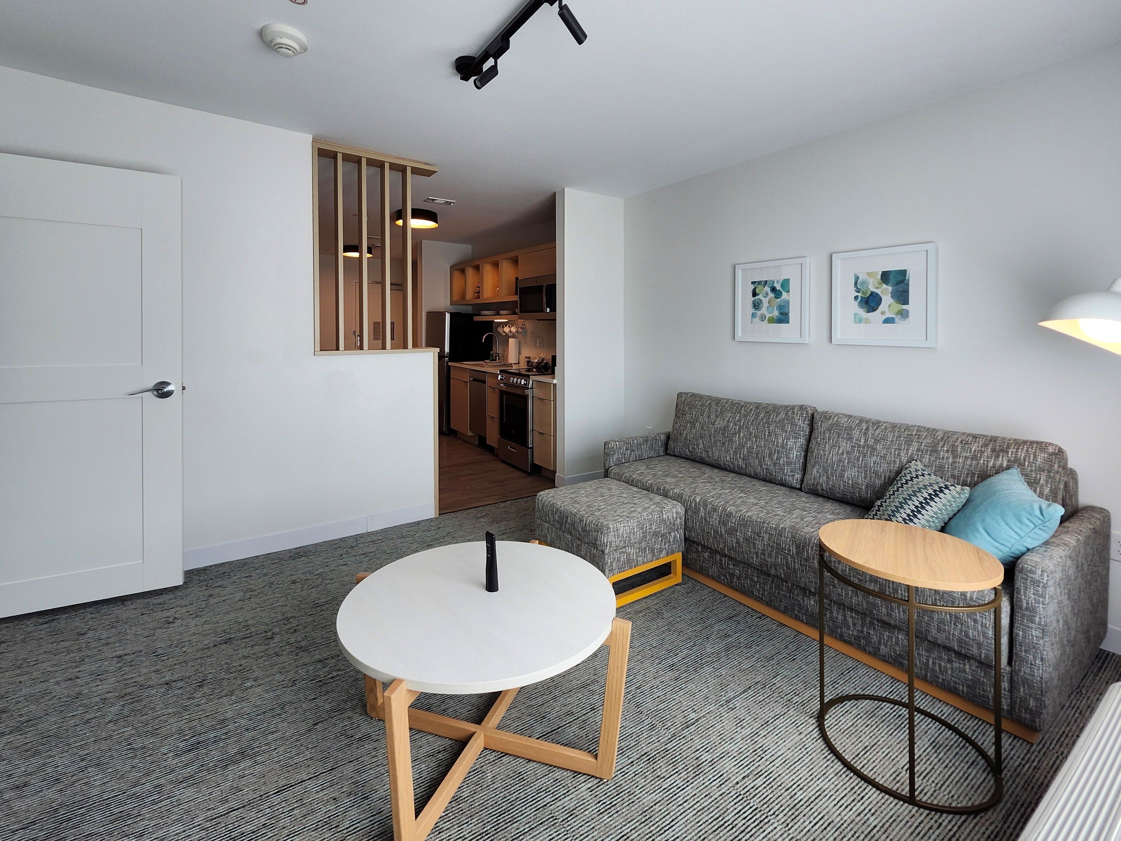 A modern living room with a grey sofa, a round wooden table, and a white table with a black object on it.