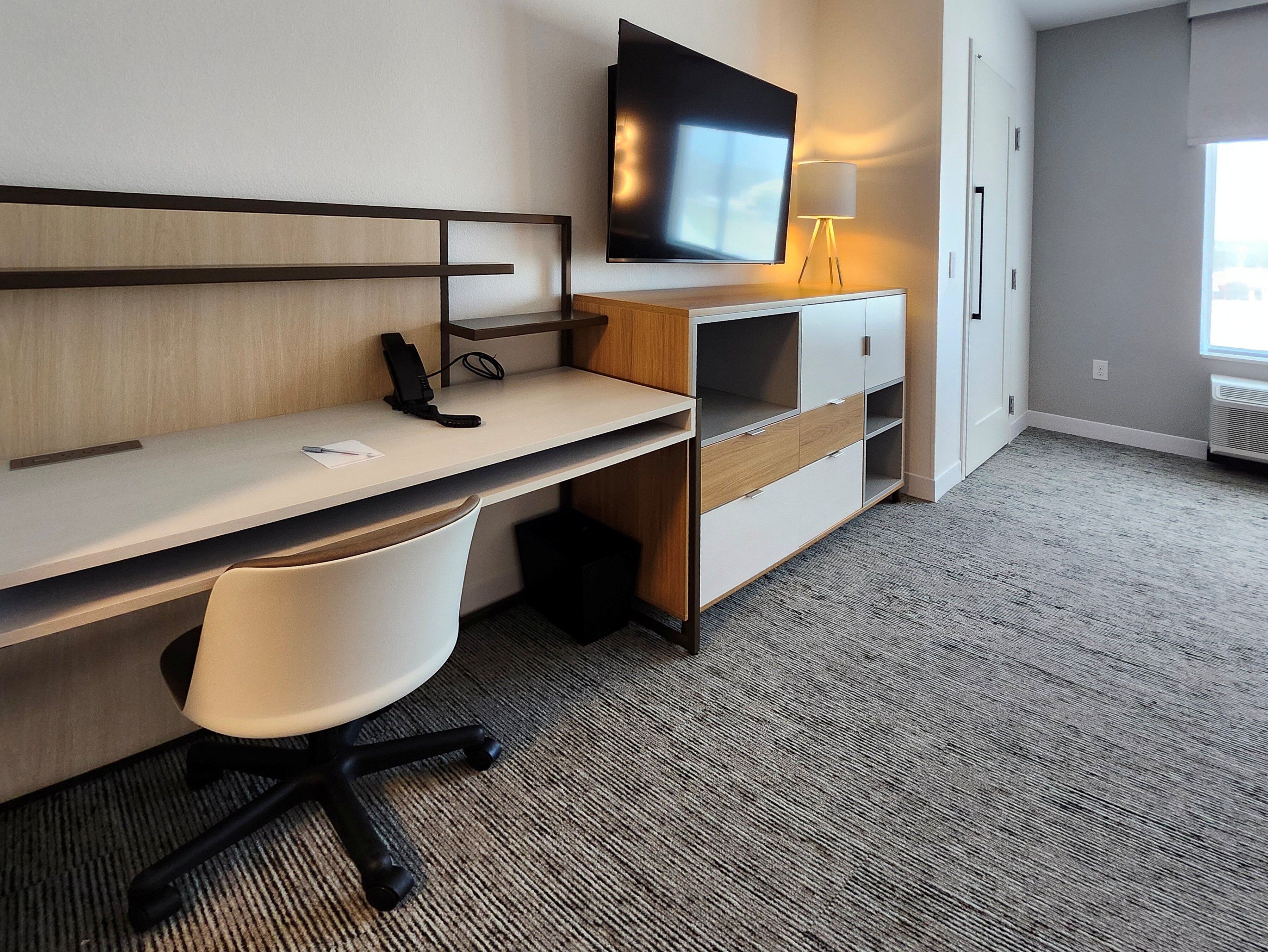 A hotel room with a desk, chair, television, lamp, and a window.