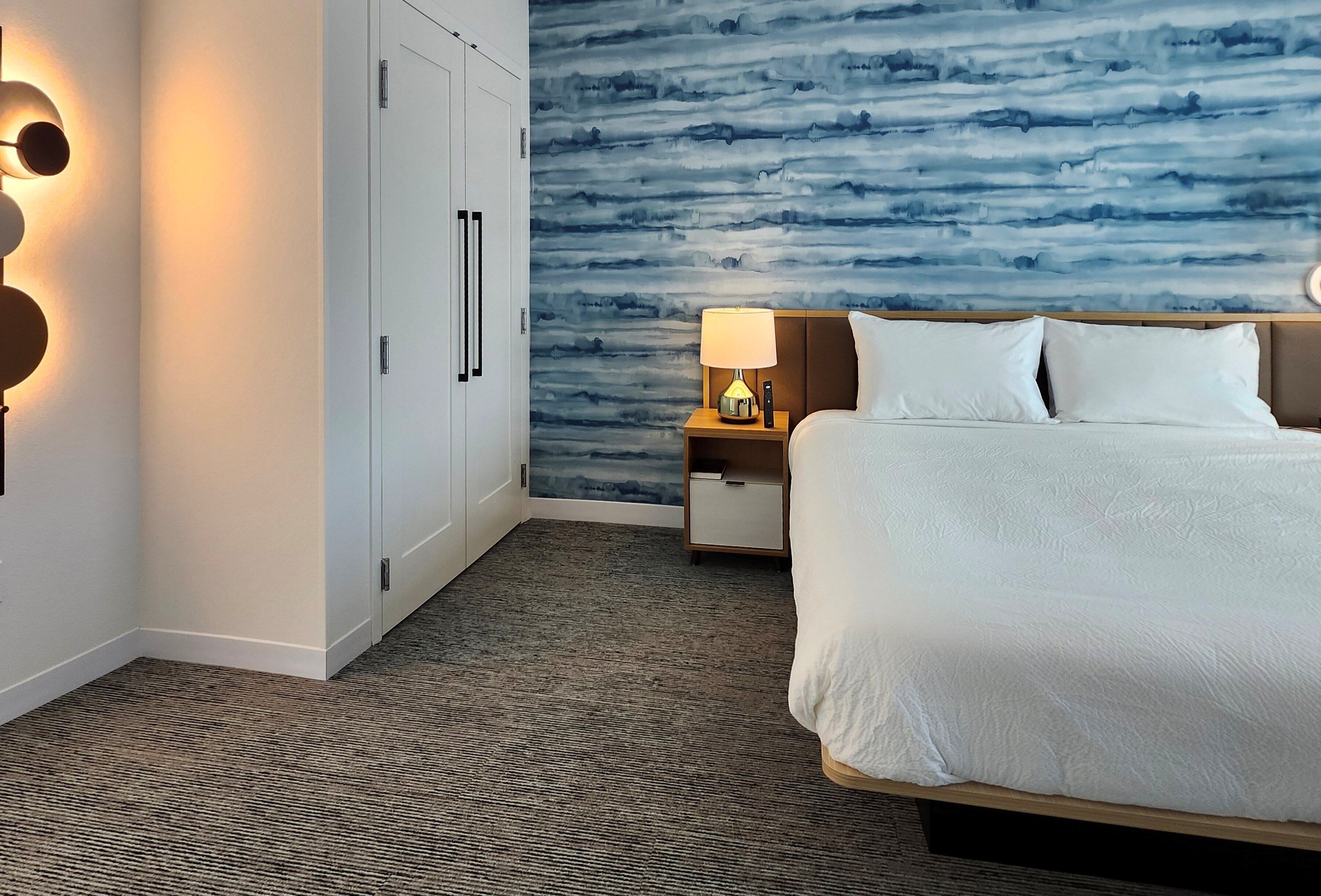 A hotel room with a bed, a nightstand, a lamp, a wall with a blue abstract design, and a wardrobe with a handle.