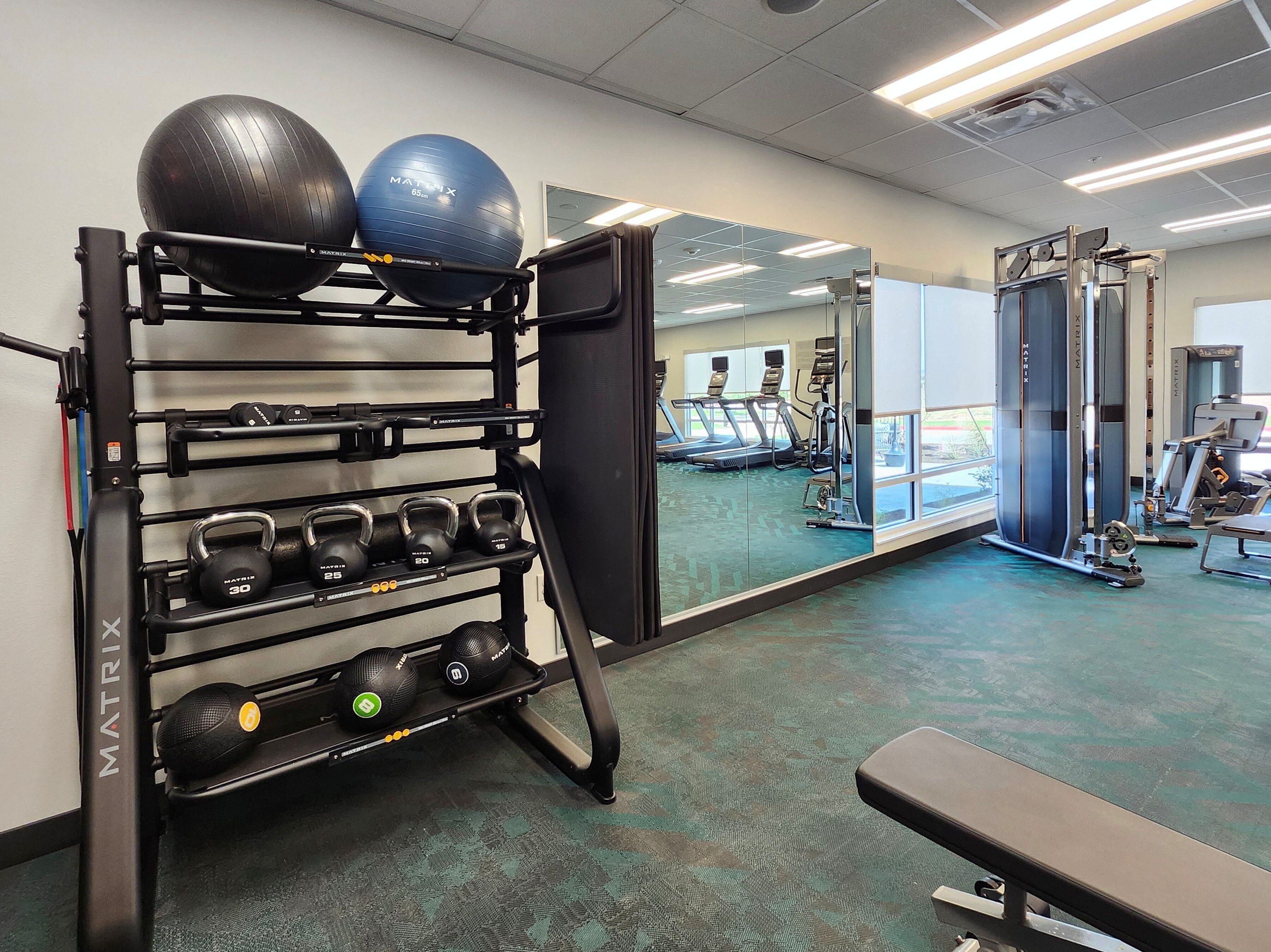 A gym with exercise equipment, including a rack with medicine balls and kettlebells, a bench, and a mirrored wall reflecting treadmills and other machines.