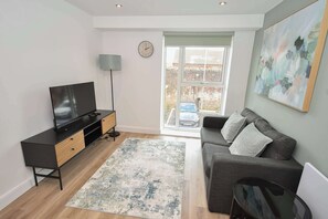 A modern living room with a flat-screen TV, a sofa, a coffee table, and a large window with a view of a parked car and a brick wall. - Bright & Contemporary 1 Bedroom Apartment in Leeds (Leeds)