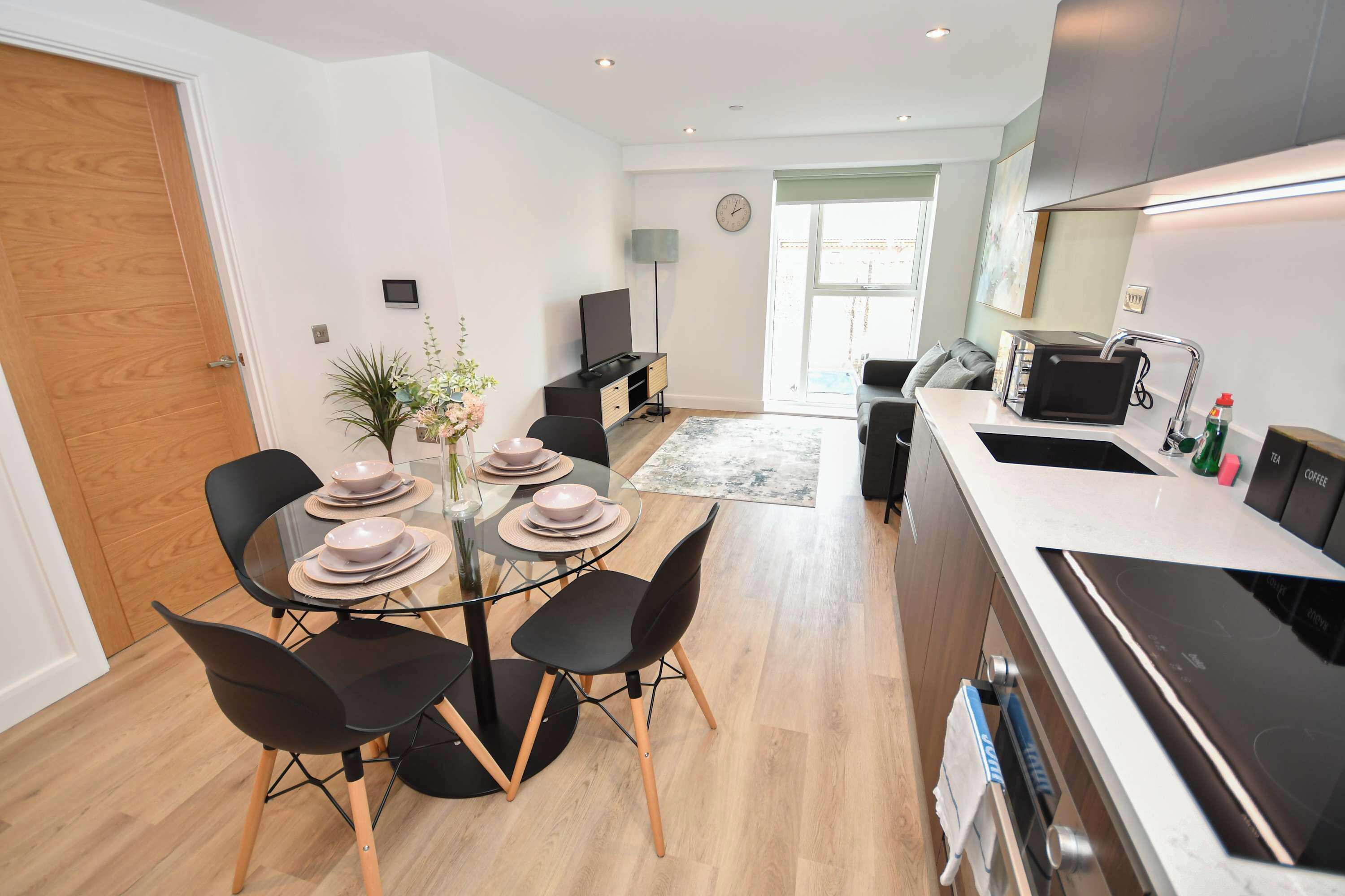 A modern kitchen with a dining area, featuring a round glass table with plates, black chairs, a microwave, and a coffee machine.