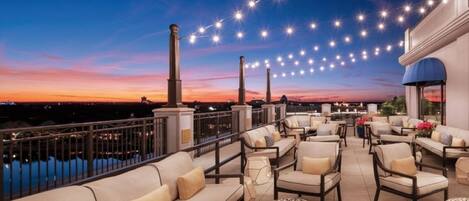Una terraza en la azotea con mobiliario exterior, luces colgantes y vistas a la ciudad al anochecer.