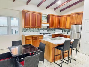 A kitchen with wooden cabinets, a stainless steel refrigerator, a built-in island with a sink, and a dining area with wicker chairs. - *Beachfront Placencia Village* off the Sidewalk! (Placencia)