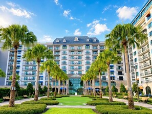 Un hotel de varios pisos con una entrada central, rodeado de palmeras y un césped cuidado.