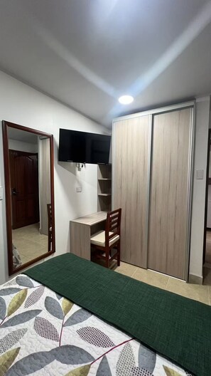 Un dormitorio con cama, escritorio, silla, armario y televisor montado en la pared.