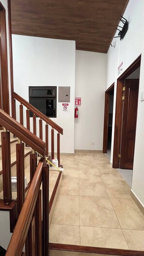 Un couloir avec un escalier en bois, un extincteur et un détecteur de fumée fixés au mur.