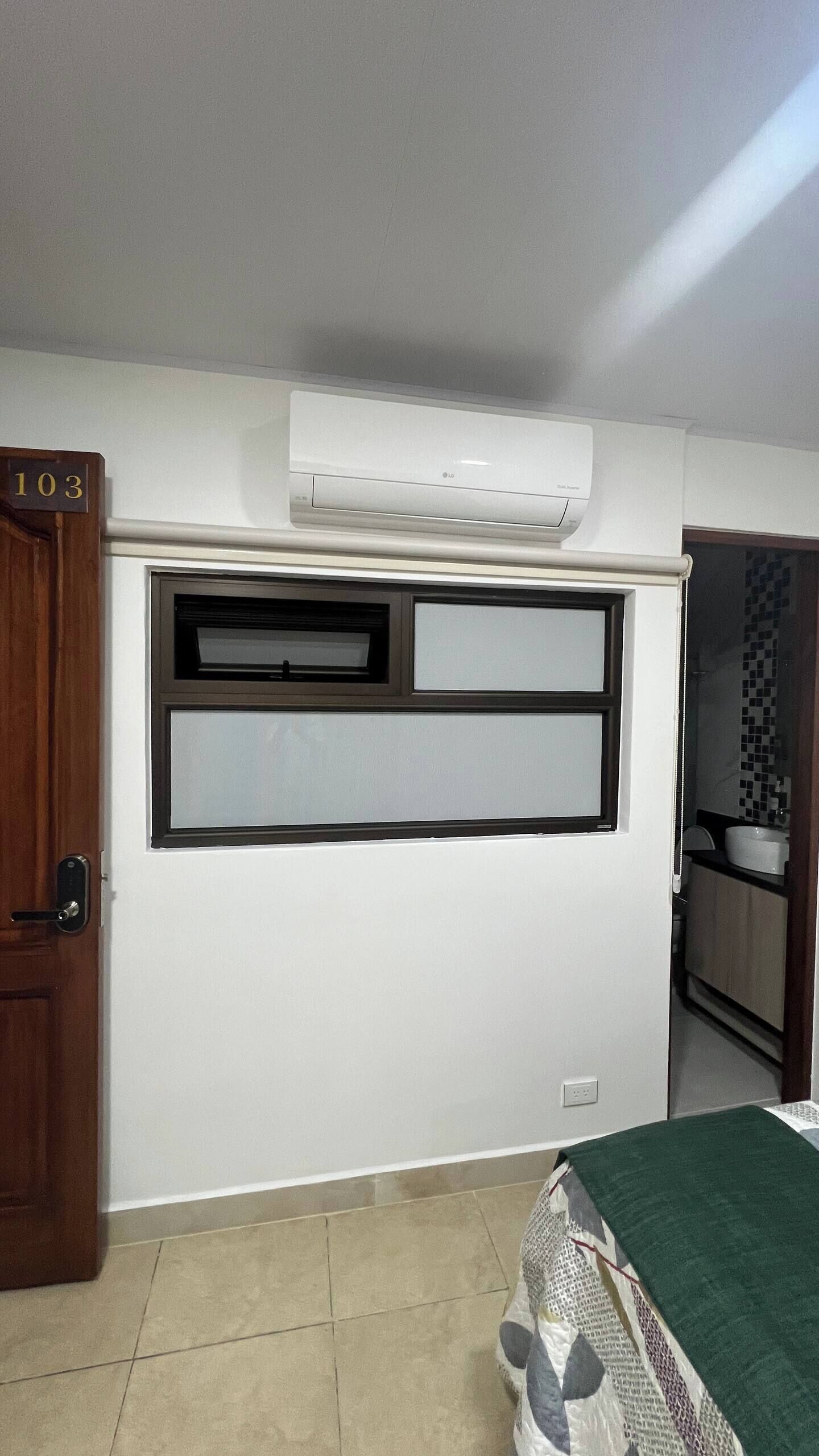 A room with a door labeled '103', a window with blinds, a ceiling-mounted air conditioner, and a bed with a green cover.