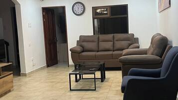 A living room with a brown sofa, a glass coffee table, and a blue armchair. There is a clock on the wall and a chandelier on the ceiling.