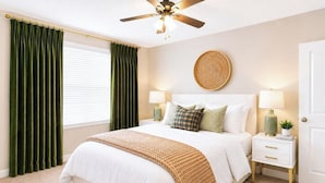 Quarto com cama, mesas de cabeceira, ventilador de teto, janela com cortinas e um objeto decorativo na parede.
