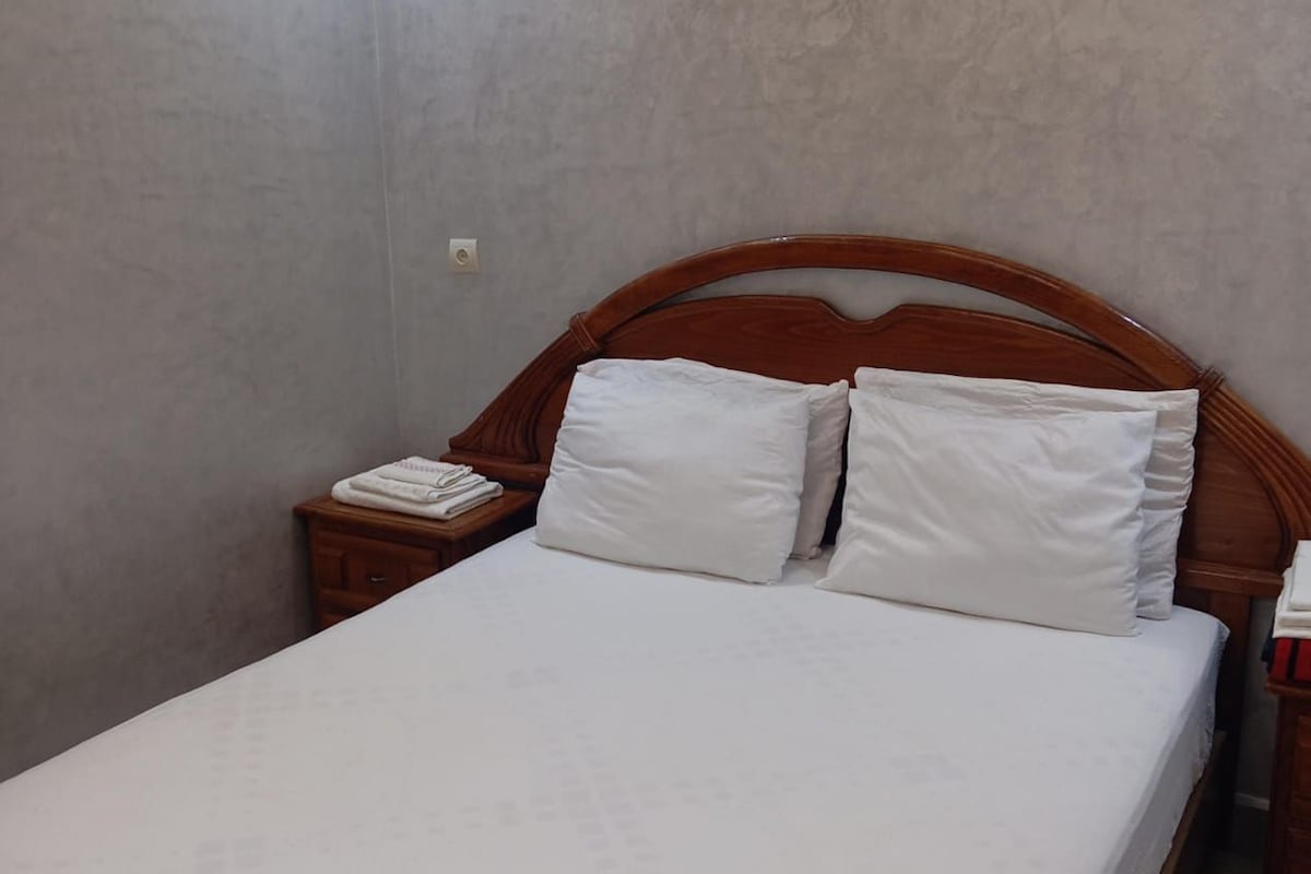 Una cama individual con cabecera de madera y ropa de cama blanca en una habitación con paredes lisas y una mesita de noche de madera.