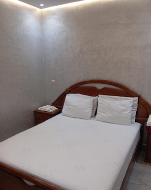 Una cama individual con cabecera de madera y ropa de cama blanca en una habitación con paredes lisas y una mesita de noche de madera.