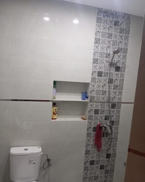 Un baño con inodoro, ducha y pared de azulejos con diseño, además de un estante con artículos de aseo.