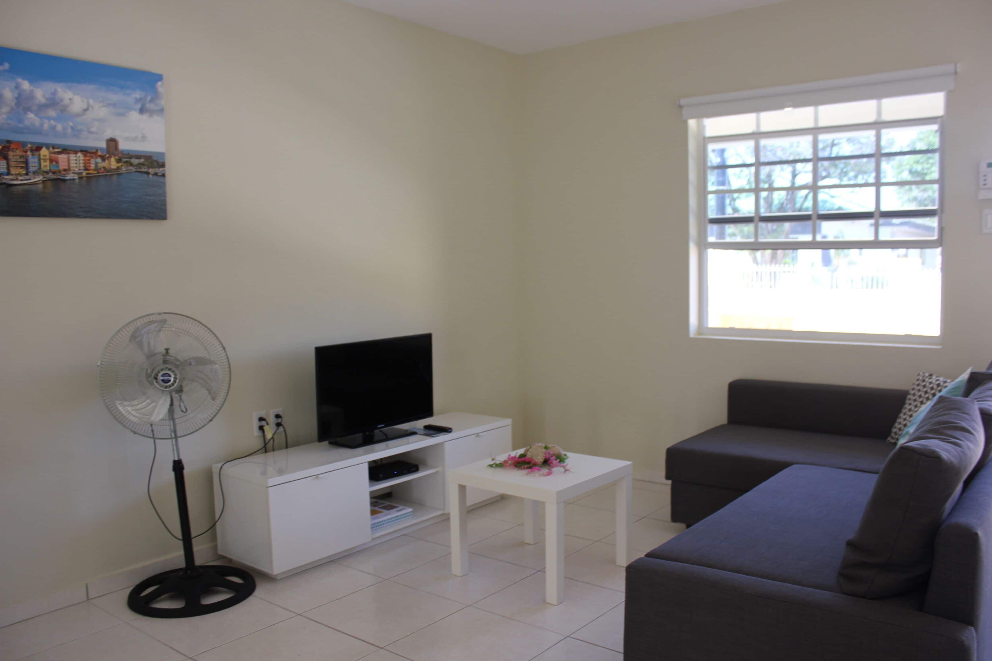 A living room with a television, a fan, a sofa, and a window with blinds.