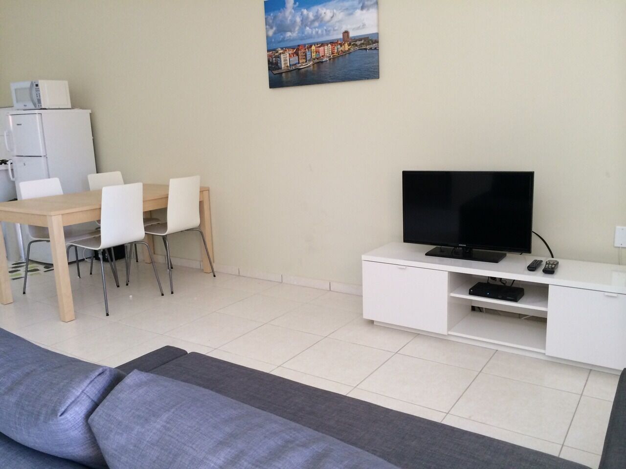 A living room with a flat-screen TV, a dining area with chairs, a sofa, and a refrigerator.