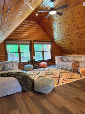 Interior acogedor de una cabaña con techo inclinado, paredes de madera, un ventanal grande, un ventilador de techo y un colcha de colores en el suelo.