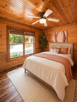 A wooden room with a bed, a ceiling fan, a window, and a plant.