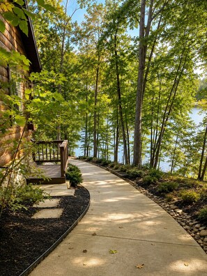 A concrete pathway leading to a wooden deck with a view of a lake surrounded by trees.