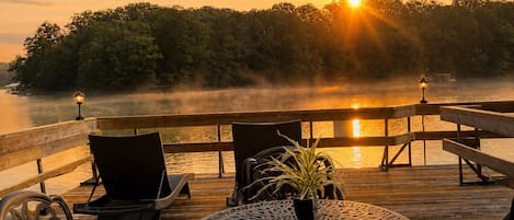Una terraza de madera con mobiliario exterior, una mesa redonda y sillas, con vista a un cuerpo de agua y un atardecer de fondo.