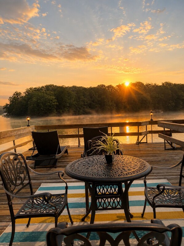 A wooden deck with outdoor furniture, a round table, and chairs, overlooking a body of water with a sunset in the background.