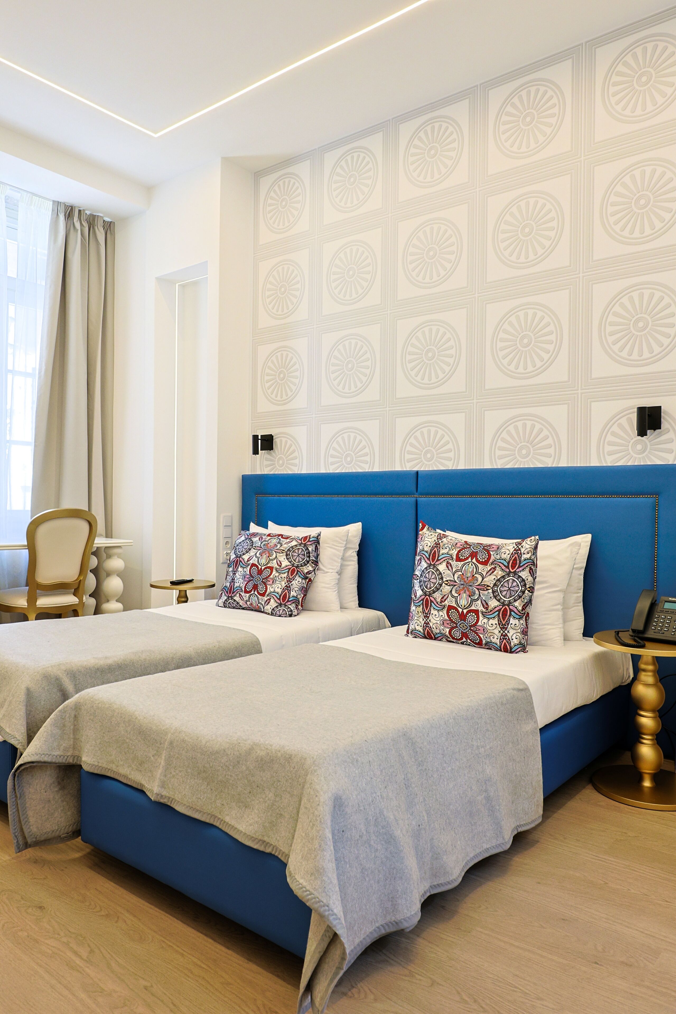 Une chambre d’hôtel avec deux lits, une tête de lit bleue, des motifs décoratifs sur les murs, une chaise, une table de chevet avec un téléphone et une fenêtre avec des rideaux.