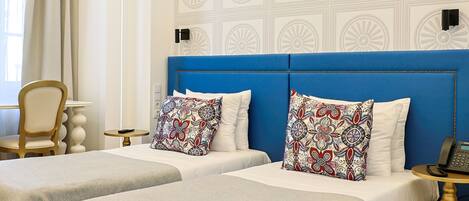 Une chambre d’hôtel avec deux lits, une tête de lit bleue, des motifs décoratifs sur les murs, une chaise, une table de chevet avec un téléphone et une fenêtre avec des rideaux.