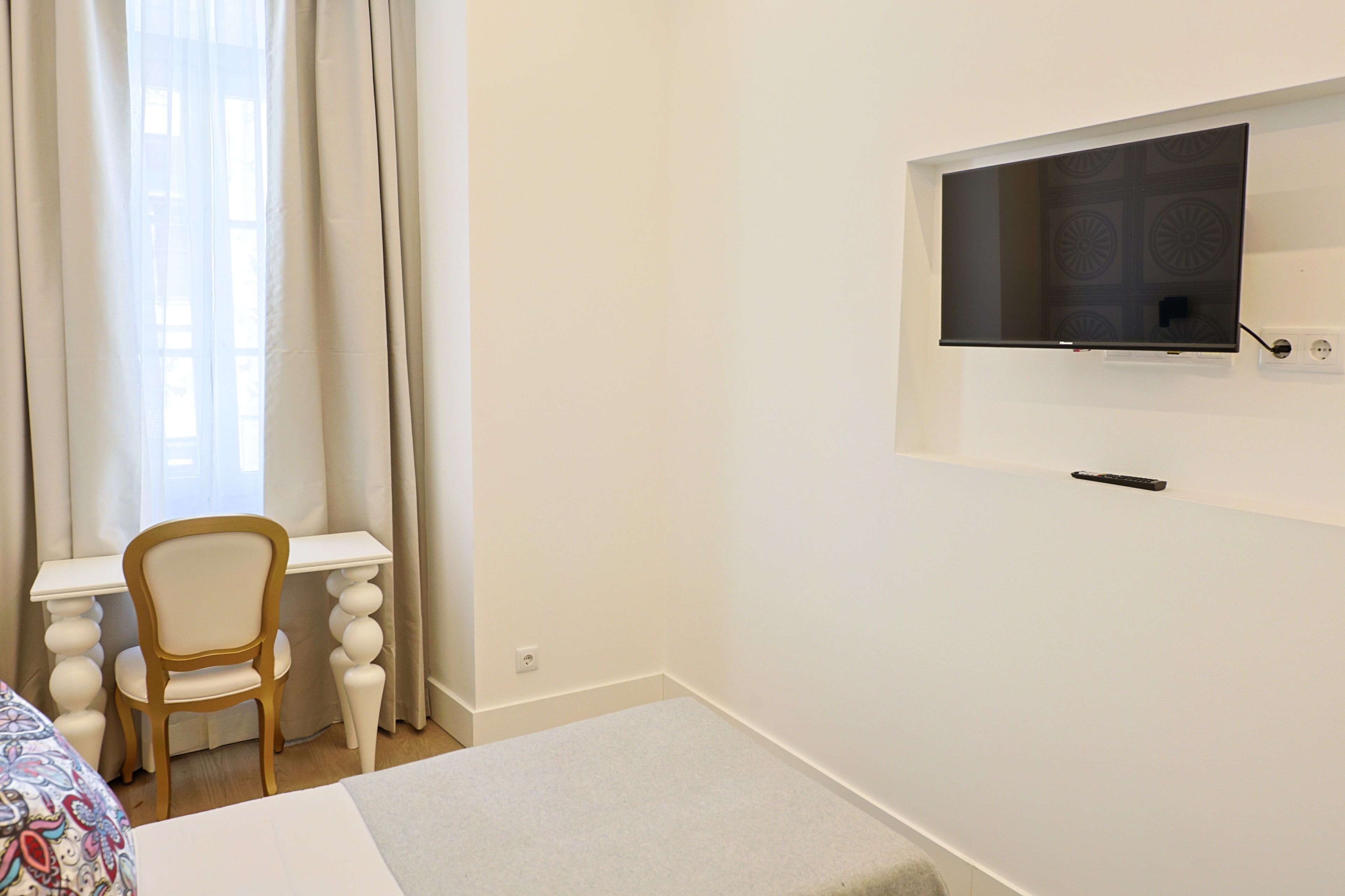A hotel room with a bed, a desk, a chair, a television mounted on the wall, and a window with curtains.