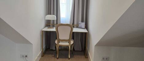 A corner room with a wooden floor, a desk with a lamp, a chair, and a window with curtains.