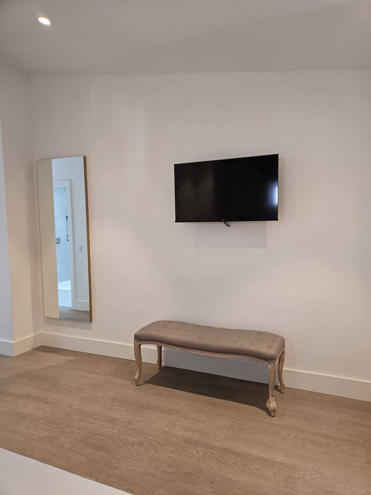A room with a flat-screen TV mounted on the wall, a bench, and a door leading to another room.