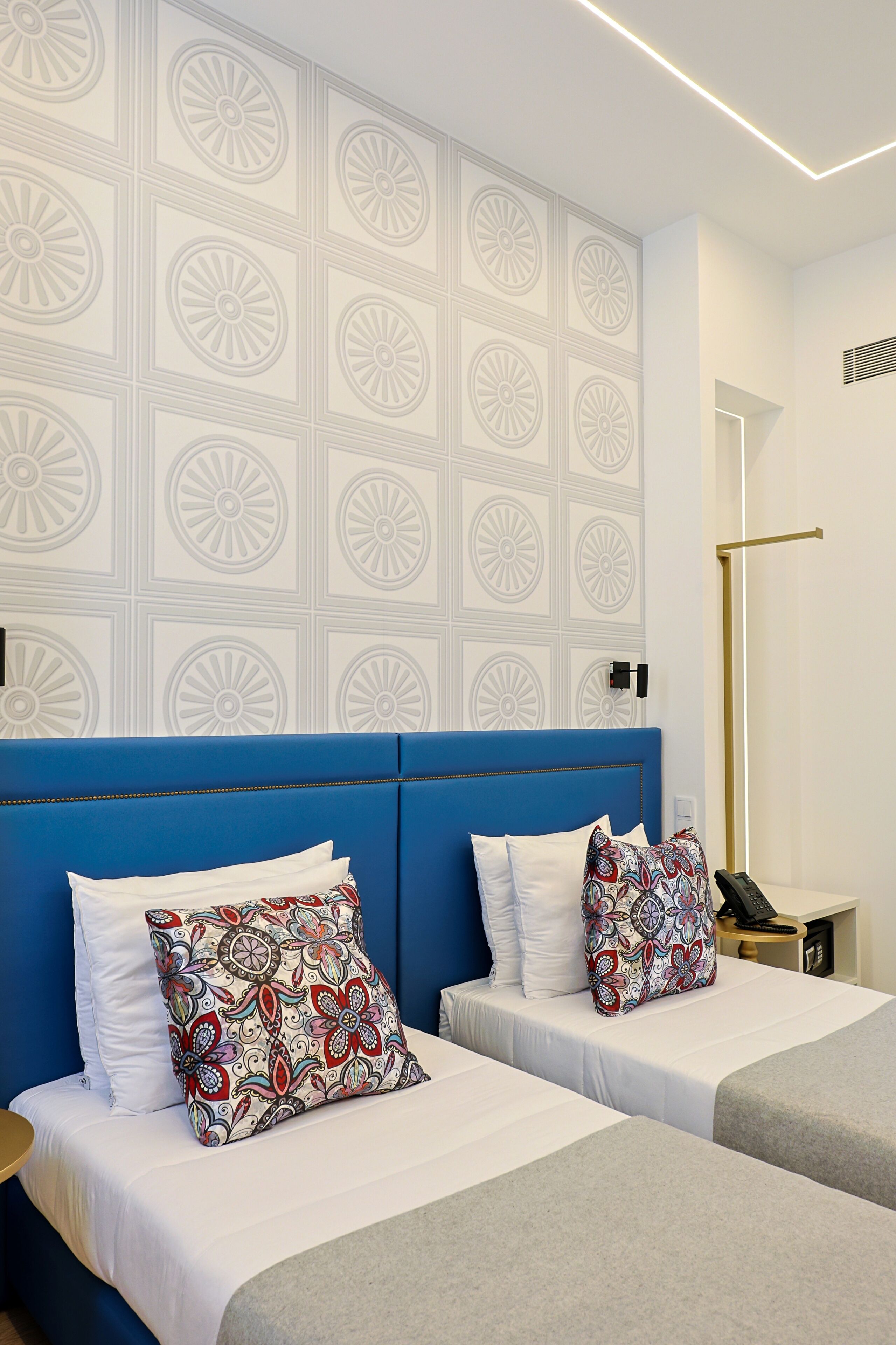 A hotel room with a blue headboard, two beds with white bedding, patterned pillows, a bedside table with a phone, and a wall with a geometric pattern.